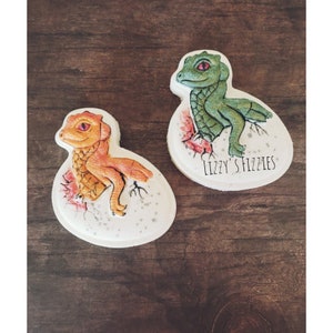 Può includere: Due biscotti di zucchero decorati, ognuno raffigurante un dinosauro neonato che esce da un uovo. Un biscotto presenta un dinosauro verde con occhi rossi, mentre l'altro biscotto presenta un dinosauro arancione con occhi rossi. I biscotti sono decorati con glassa bianca e hanno un aspetto maculato. Il testo "Lizzy's Fizzles" è scritto sul fondo di un biscotto.