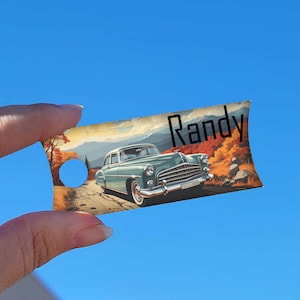 May include: A blue vintage car with the name "Randy" printed on a brown and green landscape background. The car is driving on a road with trees and mountains in the background.