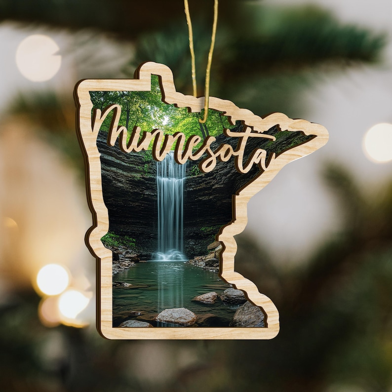 May include: Wooden ornament in the shape of the state of Minnesota, featuring a scenic waterfall image. The word "Minnesota" is scripted across the front. A gold string is attached for hanging. The ornament is likely for holiday decor.