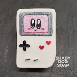 May include: White hand-painted soap shaped like a vintage handheld game console with a pink screen and a cartoon face. The console has a black plus sign button, two black lines, and two red hearts. The soap has the text "SHADY DOG SOAP" on the side.