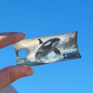 May include: A personalised tag featuring a black and white orca leaping from the water. The tag has a hole at the top for hanging and the name "LIAM" printed in blue at the bottom.