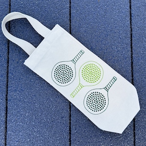 May include: White canvas tote bag with a green and black design of three paddleball rackets. The rackets have a green and white pattern.