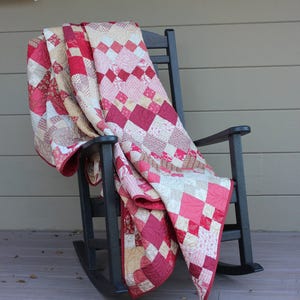 May include: A handmade quilt draped over a black rocking chair. The quilt features a patchwork design with squares in shades of red, pink, and cream. The chair is on a wooden porch, creating a cozy, inviting scene.