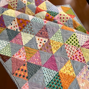 May include: A handmade quilt featuring a patchwork design of colorful triangles in various patterns and colors, including pink, orange, yellow, and green. The quilt has a gray background and a black and white striped border. The quilt is a decorative home textile.