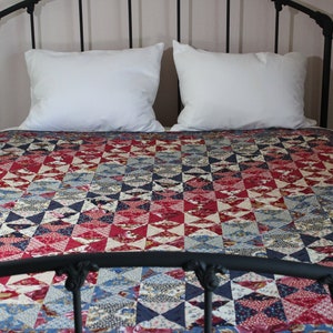 May include: A bed with a black metal frame, two white pillows, and a quilt. The quilt features a geometric pattern of red, blue, and cream-colored fabric. The bed is made up and ready for use.