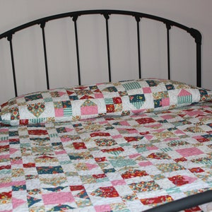 May include: A bed with a black metal frame and a patchwork quilt. The quilt features a repeating pattern of squares in shades of red, pink, blue, and green. The pillow has a matching design. The bed is in a room with a light pink wall.