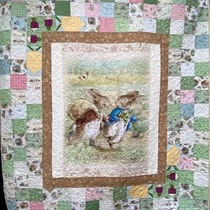 May include: A patchwork quilt featuring a central panel of Peter Rabbit and another rabbit in a garden, surrounded by a brown border and a grid of squares in pink, green, and white patterns. The central panel includes the text Peter Rabbit.