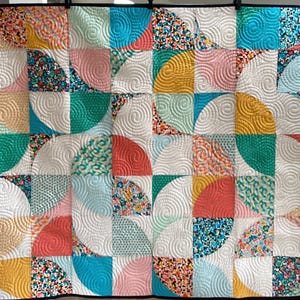May include: A colorful quilt with a patchwork design. The quilt features a pattern of quarter-circle shapes in various colors, including teal, coral, yellow, and pink, with white spiral quilting. The quilt is framed with a black border.