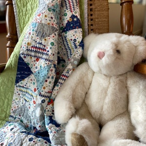 May include: A white plush teddy bear with a pink heart-shaped nose sits next to a colorful patchwork quilt. The quilt features a space-themed design with stars, planets, and rockets in shades of blue, white, and red. The quilt is draped over a wooden chair.