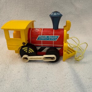 May include: A vintage toy train in yellow, red, and blue. The train features the words "TOOT-TOOT" in a white rectangle on the side. The train has black wheels with yellow accents and a yellow pull string.