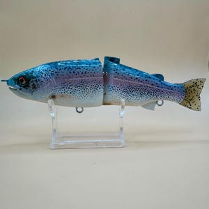 May include: A blue and purple articulated fishing lure shaped like a rainbow trout. The lure is segmented, with a hinged midsection. It has a detailed paint job with spots and a yellow-spotted tail. The lure is mounted on a clear plastic stand.