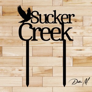 Puede incluir: Silueta negra de metal de un p&aacute;jaro con las palabras "sucker creek" en una fuente decorativa. La silueta est&aacute; unida a dos varillas de metal negro.