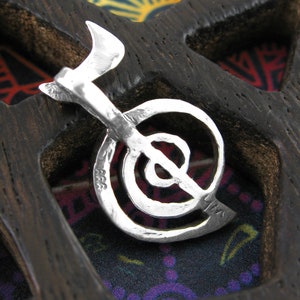 May include: A silver charm in the shape of a stylized yin yang symbol. The charm is detailed with a textured surface and is hanging from a wooden display.