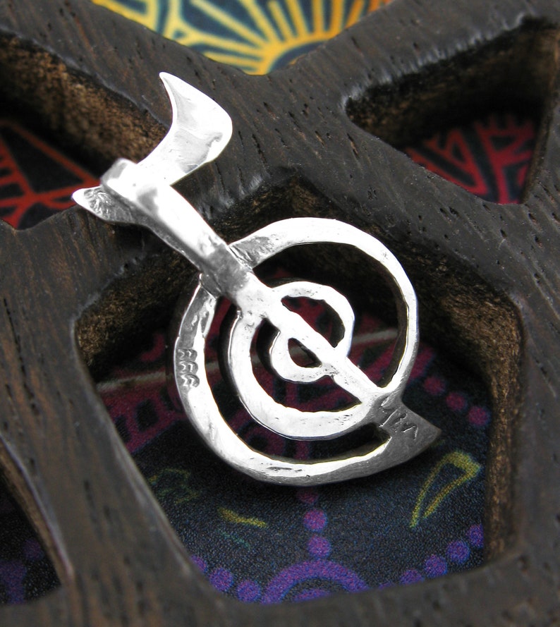 May include: A silver charm in the shape of a stylized yin yang symbol. The charm is detailed with a textured surface and is hanging from a wooden display.