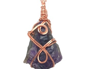 Op de afbeelding: Een ruwe amethist hanger ketting. De paarse en groene edelsteen is gewikkeld in koperdraad en hangt aan een koperen ketting. De hanger heeft een natuurlijke, onregelmatige vorm.