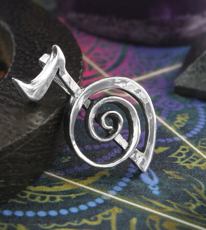 May include: Silver pendant featuring a spiral design with a small, pointed extension at the top. The pendant has a hammered texture.