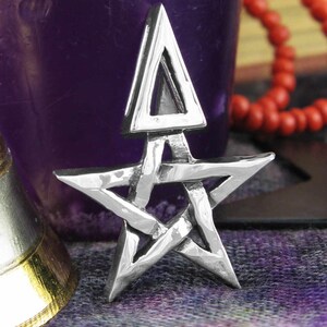 May include: A silver-colored pentagram pendant with an arrow-like design at the top. The star has a textured surface and is set against a blurred background with a purple candle holder and red beads.