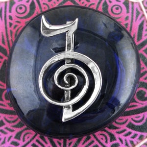 May include: A silver-colored metal symbol of a spiral with a vertical line in the center, set on a dark blue stone. The stone is round and has a smooth, polished surface.