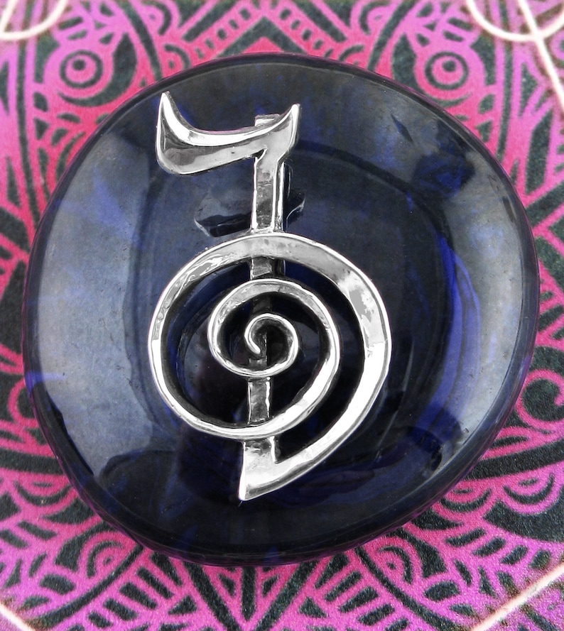 May include: A silver-colored metal symbol of a spiral with a vertical line in the center, set on a dark blue stone. The stone is round and has a smooth, polished surface.