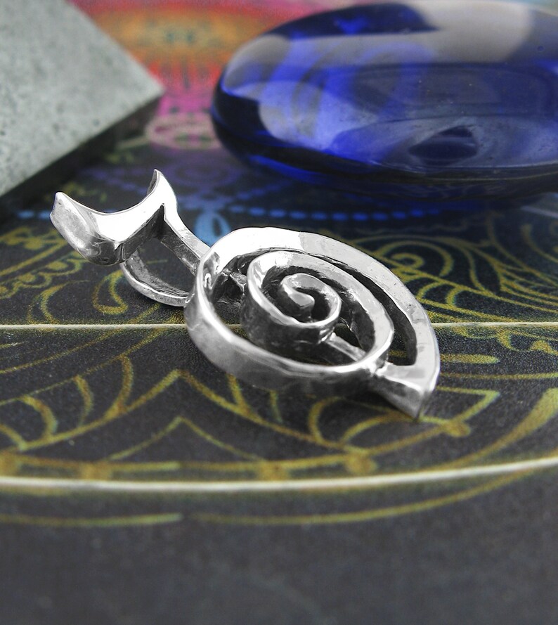 May include: A silver charm pendant in the shape of a spiral, with a smooth, polished finish. The pendant is on a dark background with a blue stone.