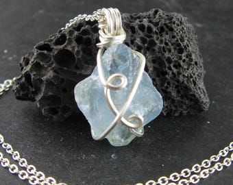 May include: A light blue, raw gemstone pendant with a silver wire wrap and chain. The gemstone is a rough, irregular shape. The silver chain is a delicate, double-strand design. The pendant is displayed on a dark, textured background.