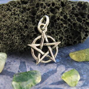 Puede incluir: Un colgante de plata con un pentagrama y un círculo alrededor de la estrella. El colgante está sobre un fondo gris oscuro.