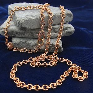 May include: A copper-colored metal chain necklace featuring round, interlocking links and a lobster claw clasp. The chain is draped in a loose, circular arrangement, showcasing the metallic finish and the consistent size of each individual link.