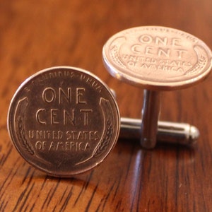 May include: A pair of cufflinks made from copper-toned US one-cent coins. The coins feature the words "One Cent" and "United States of America".