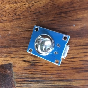 May include: A small blue circuit board with a silver sensor in the centre. The board has four holes around the edges and a small connector on the side.