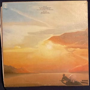 Peut inclure: Pochette de disque vinyle vintage avec une peinture de paysage de coucher de soleil, une silhouette et une sphère. Le ciel passe de l'orange au bleu, avec des titres de chansons. Un objet de collection musical.