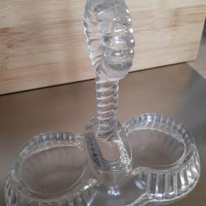 May include: Clear glass salt and pepper shaker set. The set features two circular wells connected by a clear, twisted handle. The base of the set is oval-shaped with a ribbed design around the edges. The handle is a spiral design.