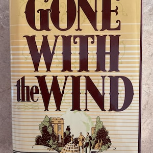 May include: A hardcover book titled "Gone With the Wind" by Margaret Mitchell. The cover is a light yellow with brown text and a detailed illustration of a mansion and figures. The title is in a large, ornate font.
