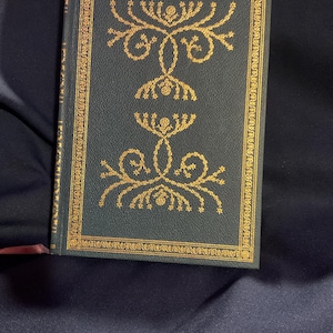 Puede incluir: Un libro de tapa dura de color verde oscuro con un borde estampado en lámina dorada y dos patrones florales simétricos en la portada. El lomo también está decorado con detalles dorados y un fino marcapáginas de cinta granate sobresale por abajo.