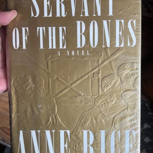 Peut inclure: Un livre doré intitulé "Servant of the Bones: A Novel" par Anne Rice. La couverture du livre présente des figures en relief et du texte en blanc, en police serif. Le livre est enveloppé dans du plastique transparent.