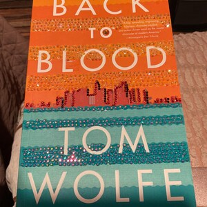 Puede incluir: Una portada de libro con el título "Back to Blood" de Tom Wolfe. La portada es naranja y azul con un diseño de ciudad y una textura brillante.