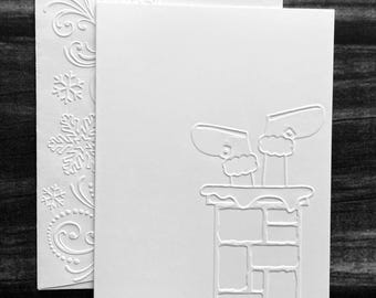May include: A white Christmas card with an embossed design of Santa's legs and boots in a chimney. The card is accompanied by a matching envelope with embossed snowflakes and swirls. The card is blank.