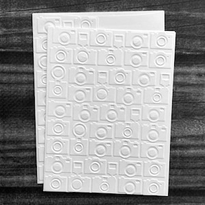 May include: Two white cards with a repeating embossed camera pattern. The cameras are detailed with a lens, viewfinder, and buttons. The cards are stacked on a dark, textured surface.