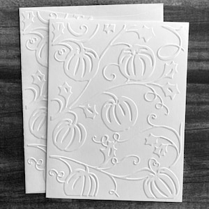May include: Two white embossed greeting cards with a pumpkin and vine design. The cards are rectangular and feature a detailed relief pattern of pumpkins, leaves, and swirling vines. The cards are stacked on top of each other.