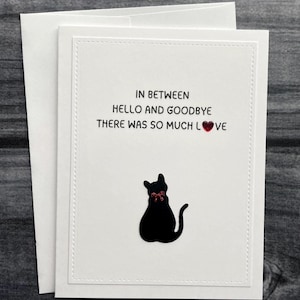 May include: A white greeting card with the text "IN BETWEEN HELLO AND GOODBYE THERE WAS SO MUCH LOVE" and a black cat silhouette with a red bow. The card is paired with a white envelope.