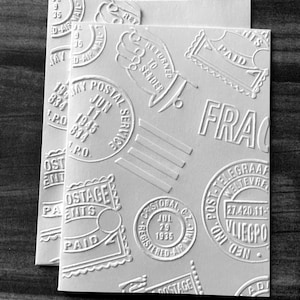 May include: Two white rectangular cards with embossed postal designs. The cards feature stamps, dates, and phrases like "PAID" and "RETURNED TO SENDER." The design is monochromatic, with a vintage aesthetic.