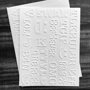 May include: White embossed sticker sheets with various text and symbols, including "SWAK", "LOL!", "TGIF", and emojis. The stickers are on a white background, with a dark, textured surface in the background.