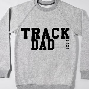 May include: A light gray sweatshirt with the words "TRACK DAD" in bold black letters. Below the text are four horizontal black lines with the numbers 1, 2, 3, and 4 on the right side. The sweatshirt has a crew neck and ribbed cuffs.