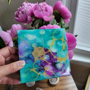 May include: Square tile with abstract art featuring teal, purple, and gold paint. The tile has a glossy finish and is perfect for adding a touch of color and texture to any space.