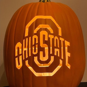 Puede incluir: Una calabaza naranja tallada con las palabras "OHIO STATE" recortadas e iluminadas desde dentro. Una decoración clásica de Halloween, perfecta para celebraciones de otoño y aficionados al fútbol americano.
