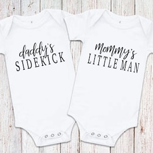 May include: Two white baby onesies with black text. One onesie says "Daddy's Sidekick" and the other says "Mommy's Little Man".