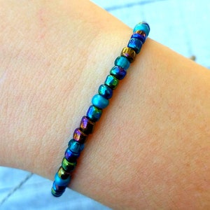 May include: A bracelet made of iridescent blue, green, and gold beads. The bracelet is on a person's wrist.