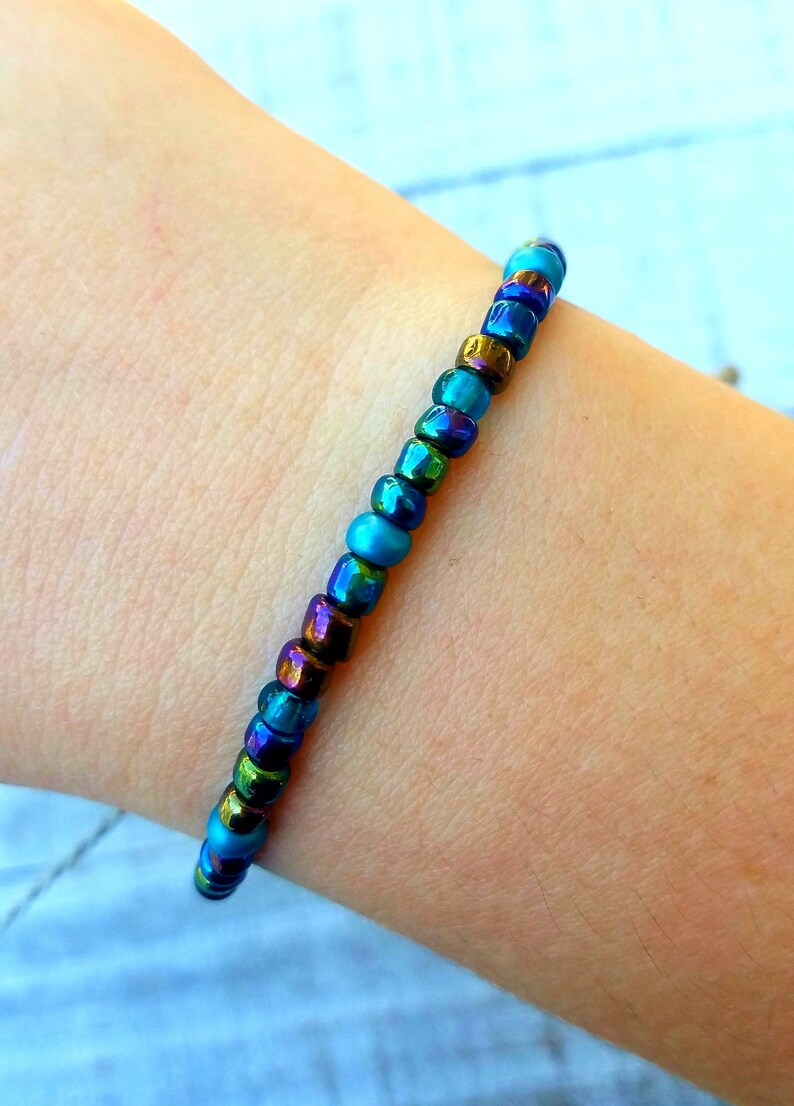 May include: A bracelet made of iridescent blue, green, and gold beads. The bracelet is on a person's wrist.