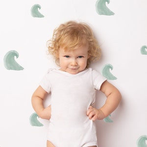 May include: A young child with blonde hair wearing a white onesie is standing in front of a white wall with green watercolour wave designs.