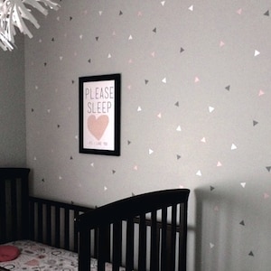 May include: A gray nursery wall with a black framed print that says "Please Sleep" with a pink heart. The wall is decorated with a pattern of small gray, pink, and white triangles.