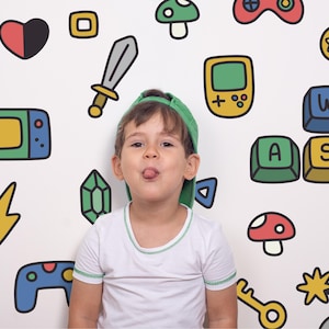 May include: A white t-shirt and a green baseball cap against a wall featuring colourful cartoon illustrations of video game controllers, a sword, a handheld console, WASD keyboard keys, hearts, mushrooms, lightning bolts, and gems.